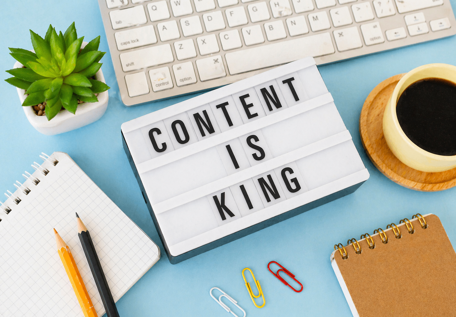 Flat lay workspace with keyboard, coffee, notebook, and lightbox displaying “Content is King” on a blue background.